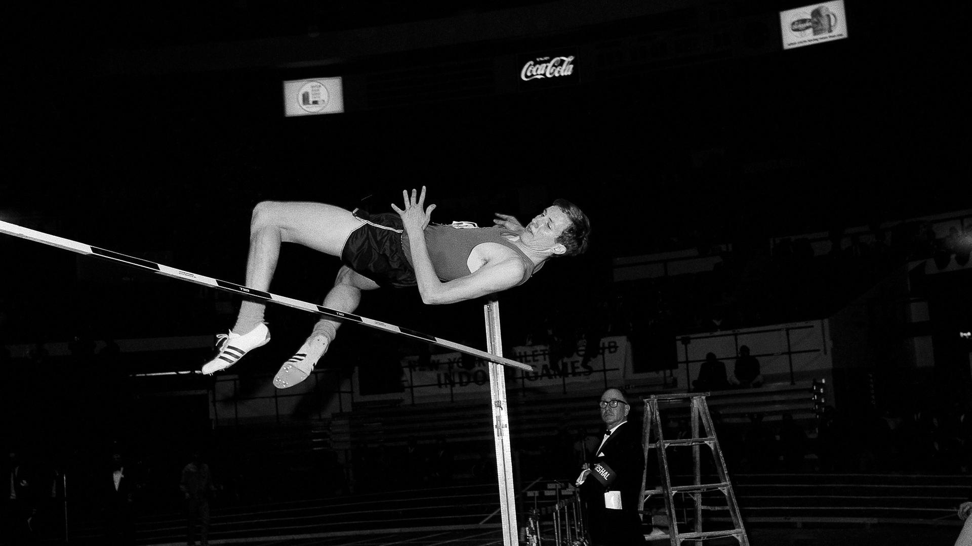 Man sieht Dick Fosbury wie er mit dem Rücken nach unten über eine Mess-Latte springt. Man sieht Dick Fosbury wie er mit dem Rücken nach unten über eine Mess-Latte springt.