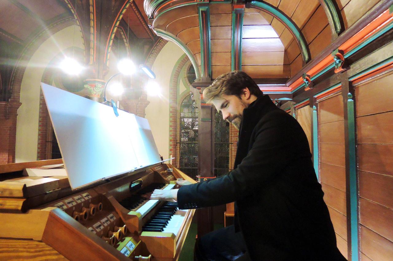 Der englische Jazz-Keyboarder Kit Downes sitzt lächelnd an einer Orgel, während er in einem dicken, schwarzen Mantel spielt. Der englische Jazz-Keyboarder Kit Downes sitzt lächelnd an einer Orgel, während er in einem dicken, schwarzen Mantel spielt.