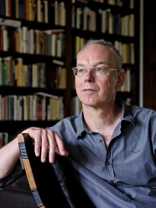 Der Autor und Journalist Günther Wessel sitzt entspannt auf einem Stuhl vor einer Bibliothek.