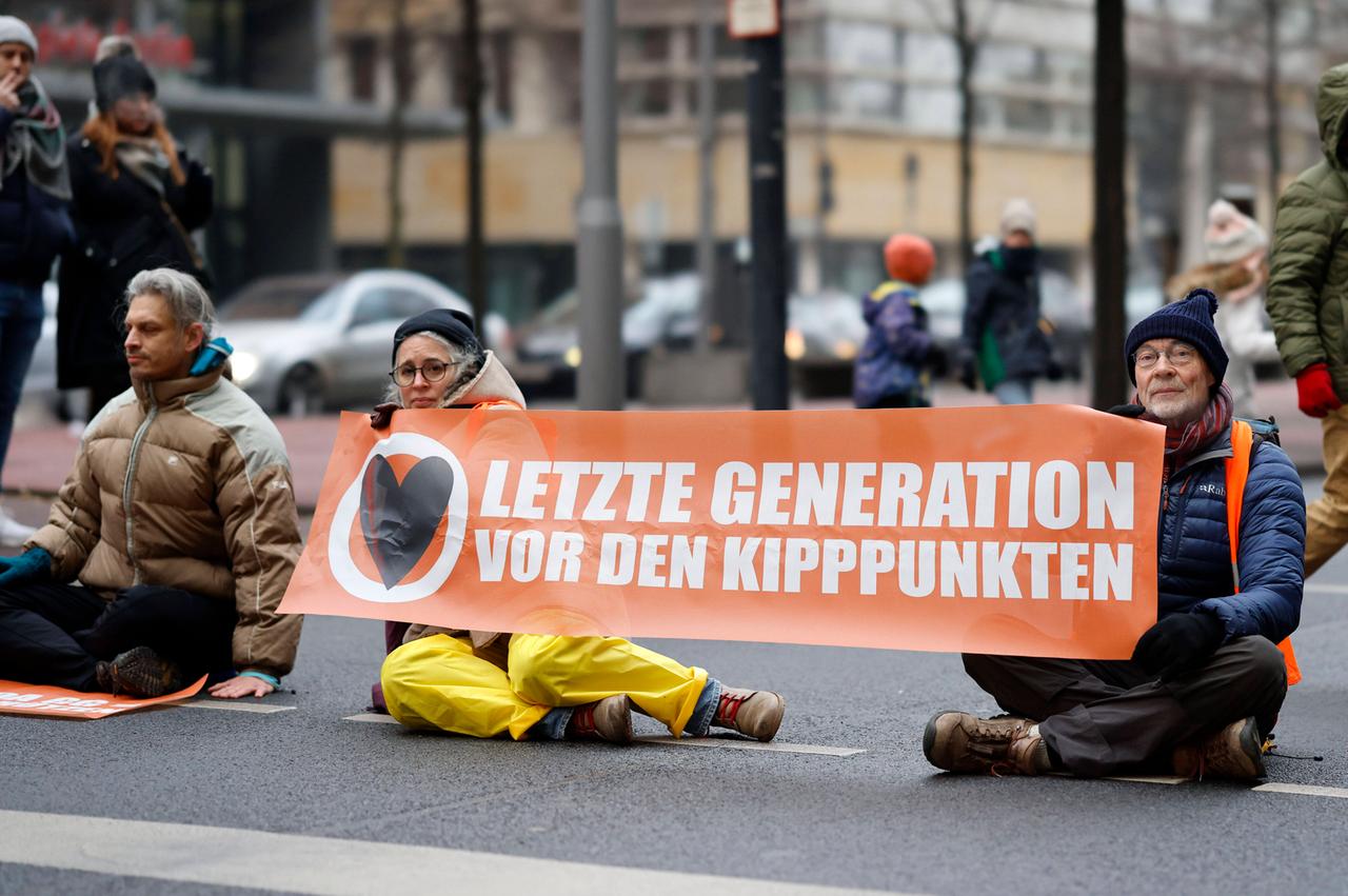 Drei Personen in Winterkleidung sitzen auf der Straße. Die Person links und die Person in der Mitte haben eine Hand flach auf der Straße. Die Person rechts hält gemeinsam mit der Person in der Mitte ein Transparent mit der Aufschrift: "Letzte Generation vor den Kipppunkten". Drei Personen in Winterkleidung sitzen auf der Straße. Die Person links und die Person in der Mitte haben eine Hand flach auf der Straße. Die Person rechts hält gemeinsam mit der Person in der Mitte ein Transparent mit der Aufschrift: "Letzte Generation vor den Kipppunkten".
