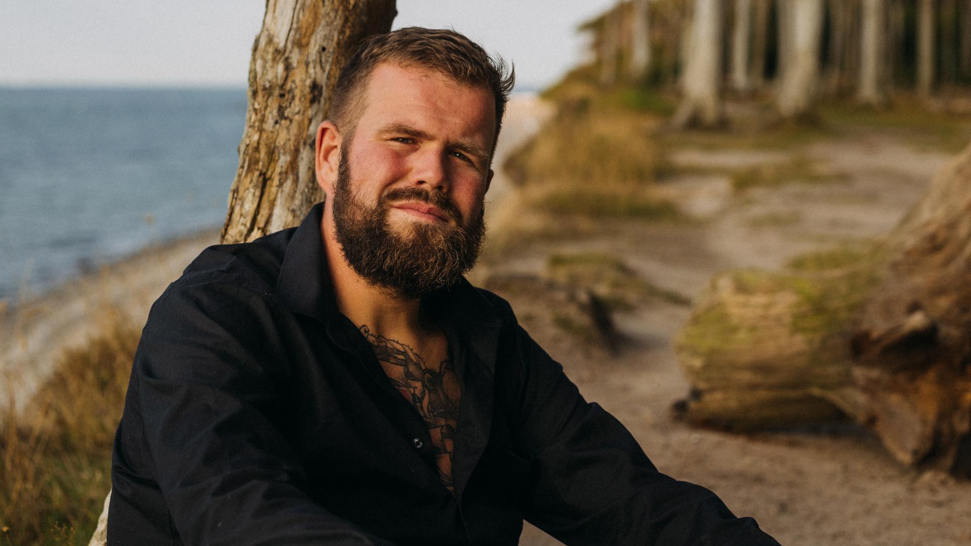 Jan Gorkow alias Monchi, Sänger von „Feine Sahne Fischfilet“, lehnt an einem Strand an einem abgestorbenen Baumstamm und blickt in die Kamera. Jan Gorkow alias Monchi, Sänger von „Feine Sahne Fischfilet“, lehnt an einem Strand an einem abgestorbenen Baumstamm und blickt in die Kamera.