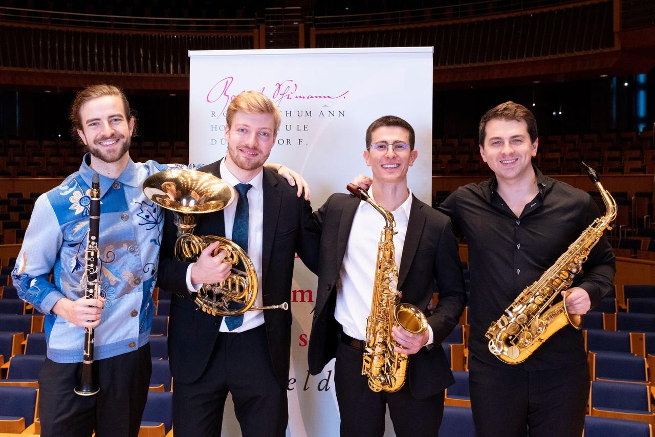 Vier junge Männer stehen neben dem Banner eines Wettbewerbes und lachen mit ihren Instrumenten in den Armen in die Kamera Vier junge Männer stehen neben dem Banner eines Wettbewerbes und lachen mit ihren Instrumenten in den Armen in die Kamera