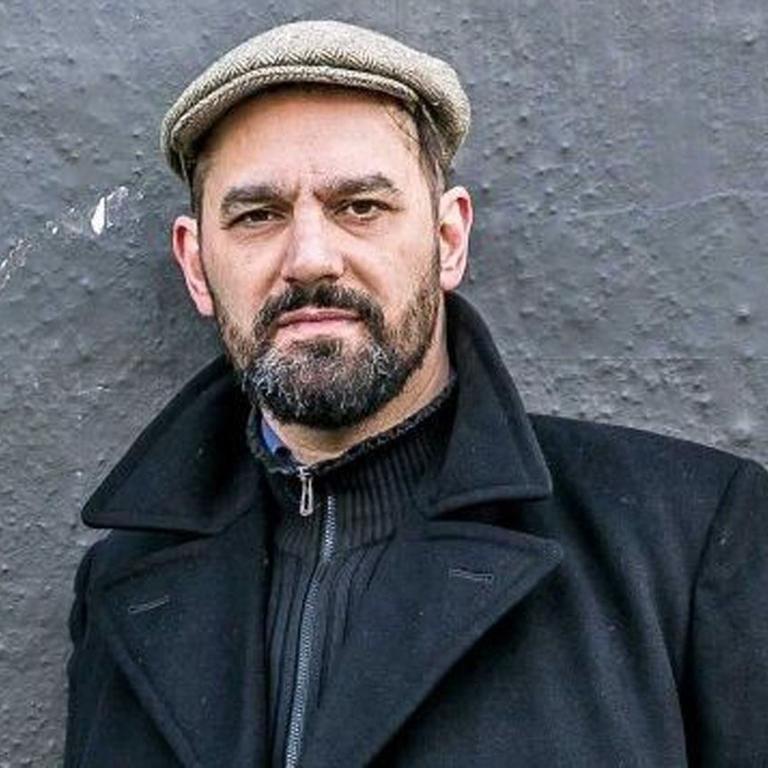 Houssam Hamade steht mit Mantel und Baskenmütze vor einer Wand.