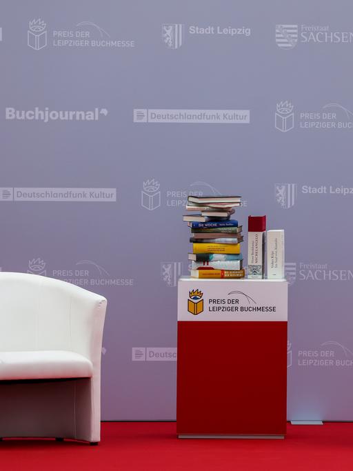 Zwei weiße Sessel und ein großer Bücherstapel auf einem Tisch stehen auf einer Bühne. Auf dem Tisch ist das Logo der Leipziger Buchmesse zu sehen, ein Auge, das auf ein geöffnetes Buch blickt.