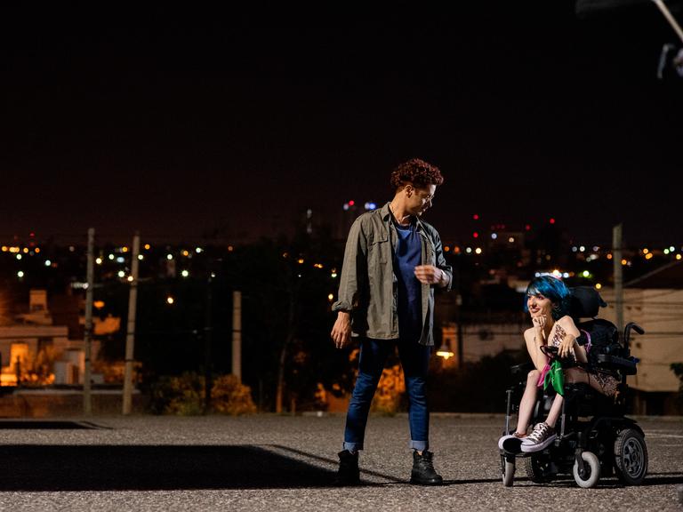 Nachtaufnahme, im Hintergrund eine beleuchtete Stadt. Am rechten Bildrand sitze die Hauptdarstellerin Juana in ihrem Rollstuhl und lacht. Vor ihr steht ein junger Mann. Sie unterhalten sich. 