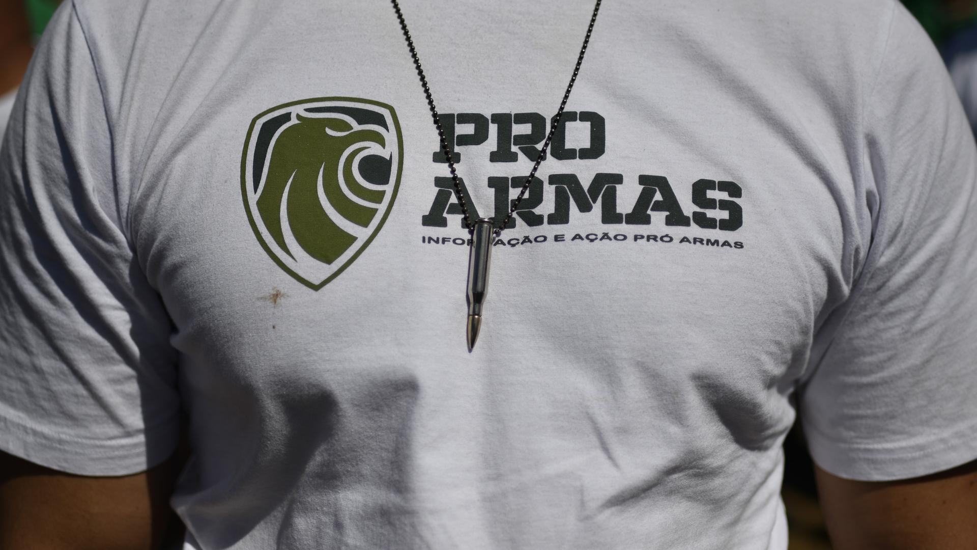 Nahaufnahme des Brustbereichs eines durchtrainierten Mannes. Er trägt ein weißes T-Shirt mit der Aufschrift "Pro Armas" und eine Kette mit einer Patrone an einem Lederband. Nahaufnahme des Brustbereichs eines durchtrainierten Mannes. Er trägt ein weißes T-Shirt mit der Aufschrift "Pro Armas" und eine Kette mit einer Patrone an einem Lederband.