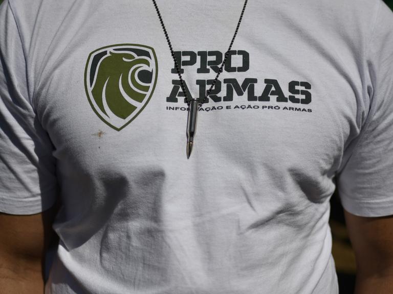 Nahaufnahme des Brustbereichs eines durchtrainierten Mannes. Er trägt ein weißes T-Shirt mit der Aufschrift "Pro Armas" und eine Kette mit einer Patrone an einem Lederband.