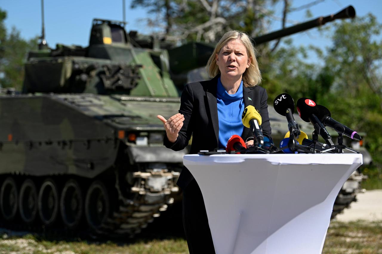 Eine blonde Frau steht im Freien an einem Pult mit Mikrofonen. Im Hintergrund ein Panzer.