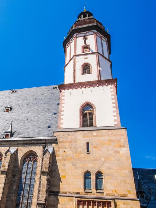 Blick von unten in die Höhe zur Turmspitze der Thomaskirche in Leipzig mit hellblauem Himmel.