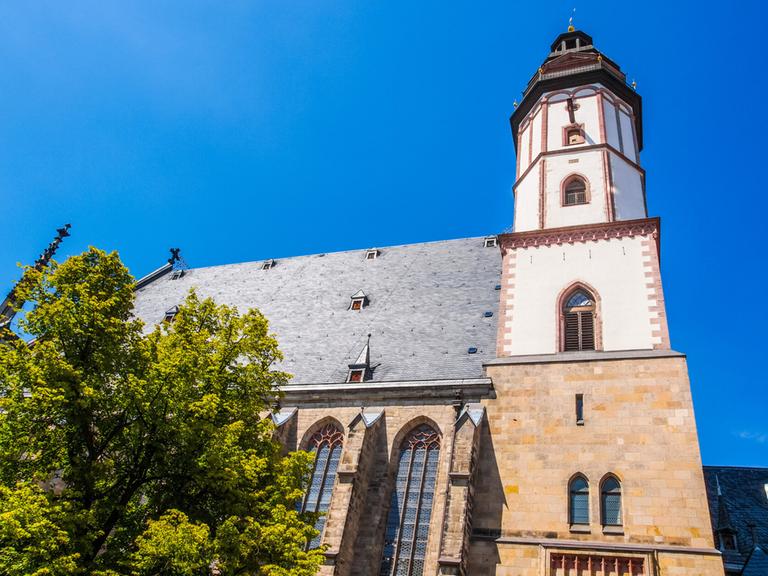 Blick von unten in die Höhe zur Turmspitze der Thomaskirche in Leipzig mit hellblauem Himmel.