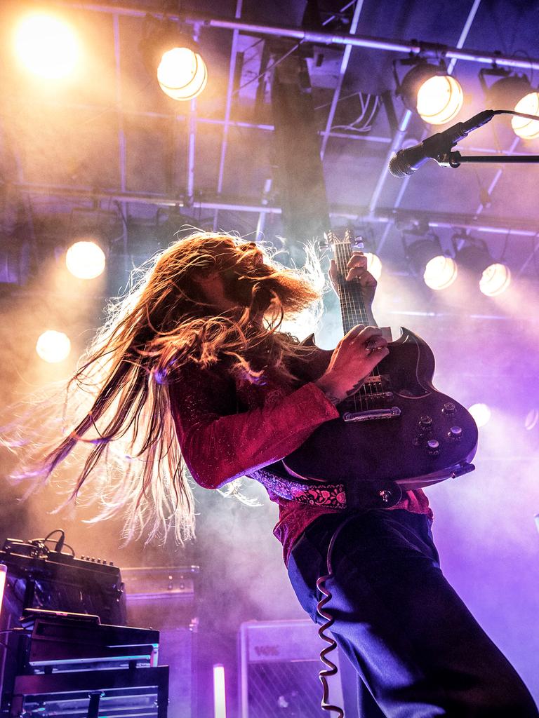 Ein Mann steht auf einer Bühne, er hälte eine E-Gitarre in der Hand und sein langes Haar schüttelt er beim Headbangen. 