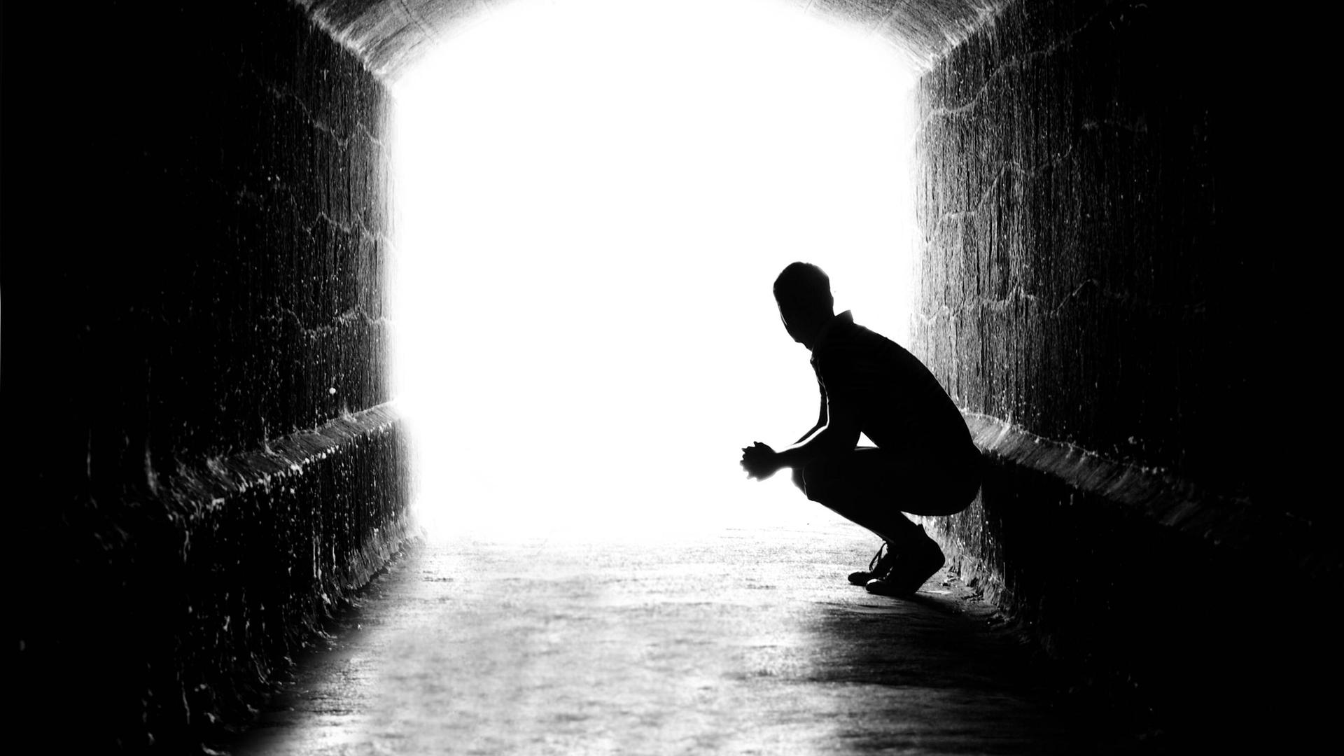 Schattenriss eines Mannes, der in einem Tunnel an der Wand hockt und in Richtung Licht blickt. Schattenriss eines Mannes, der in einem Tunnel an der Wand hockt und in Richtung Licht blickt.