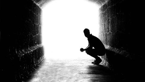 Schattenriss eines Mannes, der in einem Tunnel an der Wand hockt und in Richtung Licht blickt.
