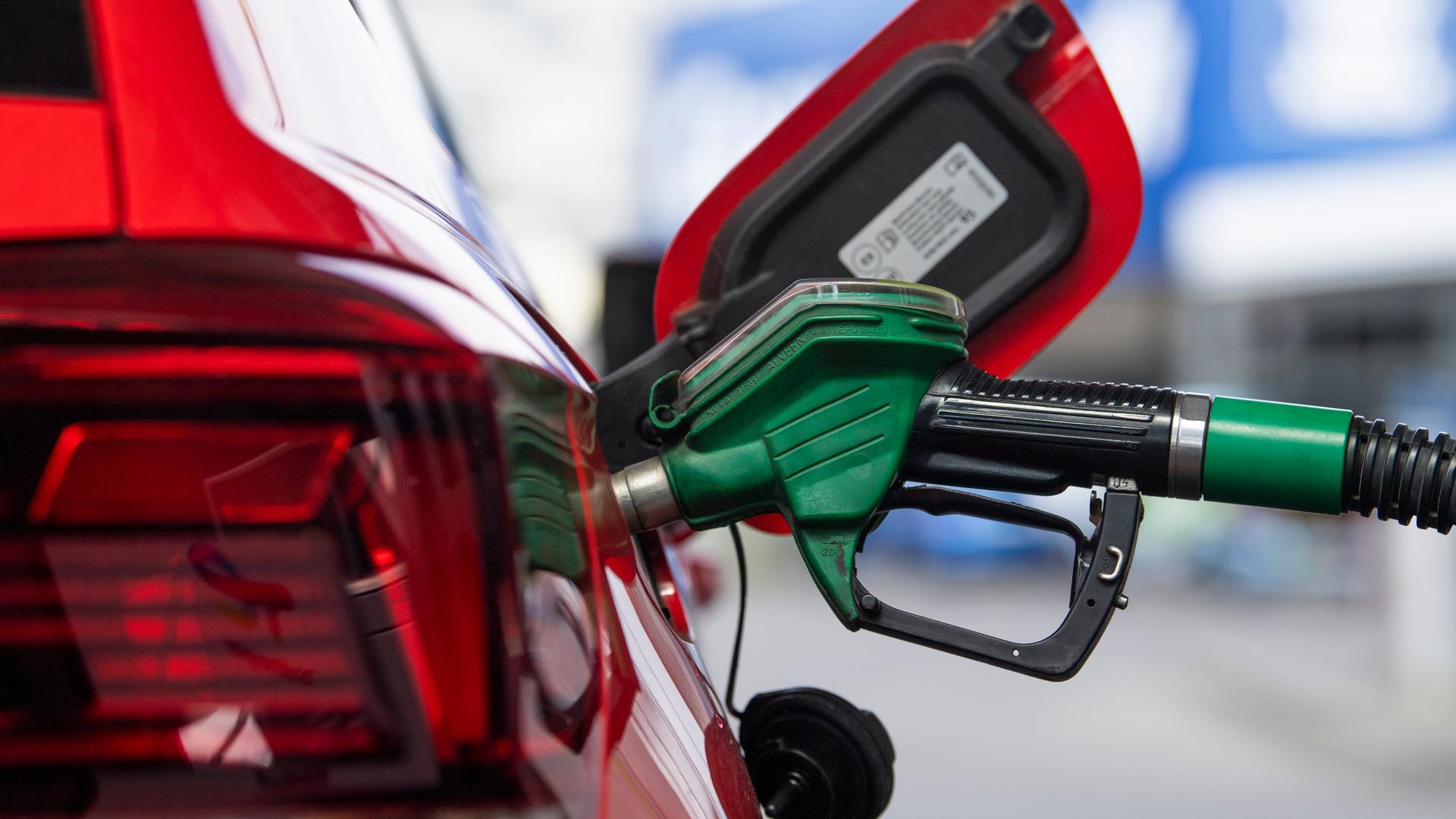 Ein grüner Einfüllstutzen steckt in der Tanköffnung eines roten Autos. Im Hintergrund unscharf das Tankstellengebäude. Ein grüner Einfüllstutzen steckt in der Tanköffnung eines roten Autos. Im Hintergrund unscharf das Tankstellengebäude.