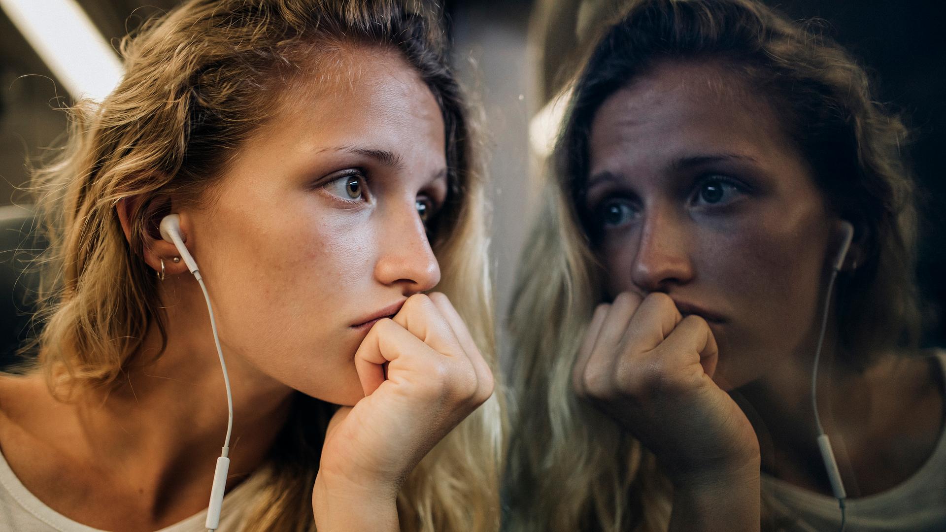 Eine junge Frau mit blonden Locken sitzt mit Kopfhörern am Zugfenster und schaut traurig aus dem Fenster, in dem sich ihr Gesicht spiegelt.
