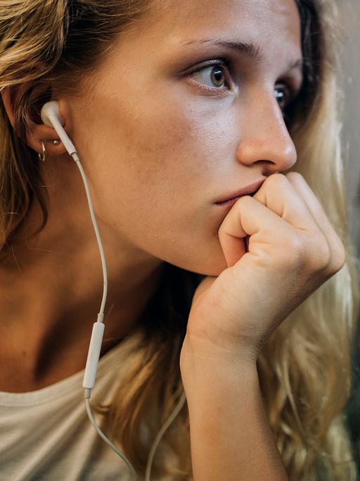 Eine junge Frau mit blonden Locken sitzt mit Kopfhörern am Zugfenster und schaut traurig aus dem Fenster, in dem sich ihr Gesicht spiegelt.
