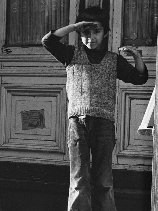 Ein türkischer Junge in Berlin-Kreuzberg 1976 steht vor einer Haustür und hält seine rechte Hand als Schutz gegen die Sonne über die Augen.