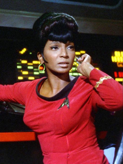Nichelle Nichols als Lieutenant Uhura in der Serie "Raumschiff Enterprise" sitzt in einer roten Uniform vor einem Schaltpult mit bunten Lichtern.
