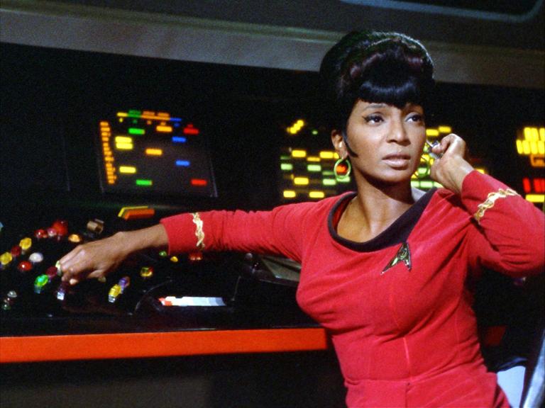 Nichelle Nichols als Lieutenant Uhura in der Serie "Raumschiff Enterprise" sitzt in einer roten Uniform vor einem Schaltpult mit bunten Lichtern.