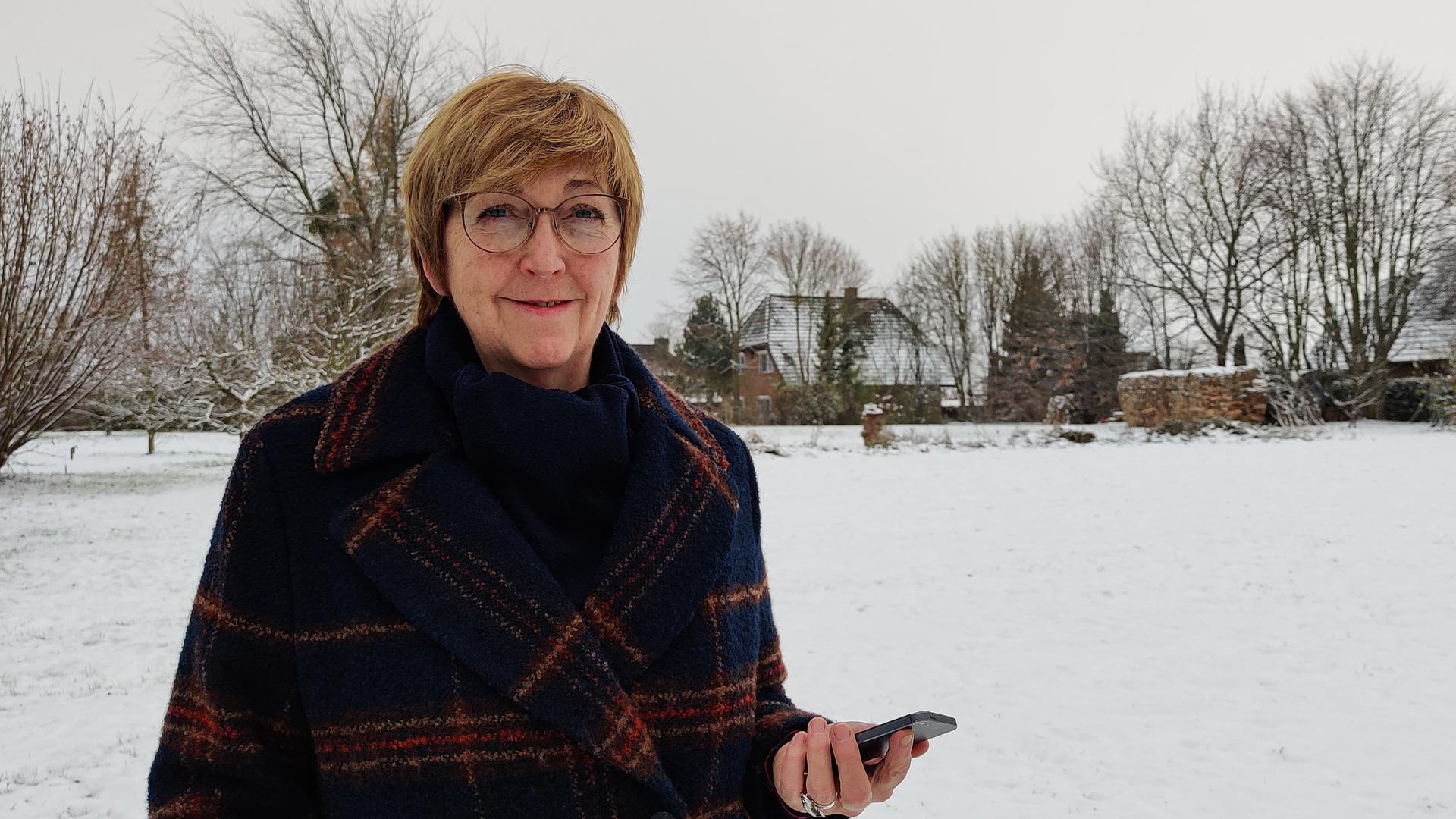 Elke Vespermann steht mit ihrem Smartphone in der Hand in einer verschneiten Landschaft. Sie trägt einen Mantel, Brille und schaut freundlich in die Kamera