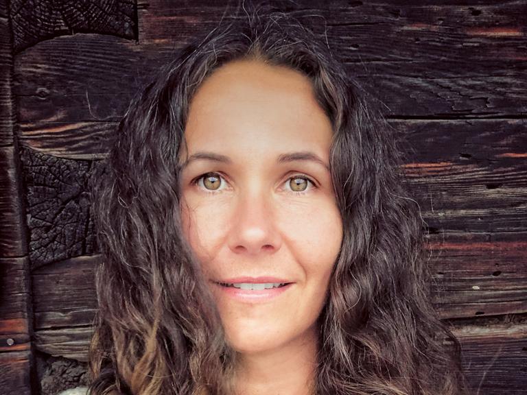 Autorin Sonja Koppitz grinst in die Kamera. Sie steht vor einer rustikalen Holzwand.