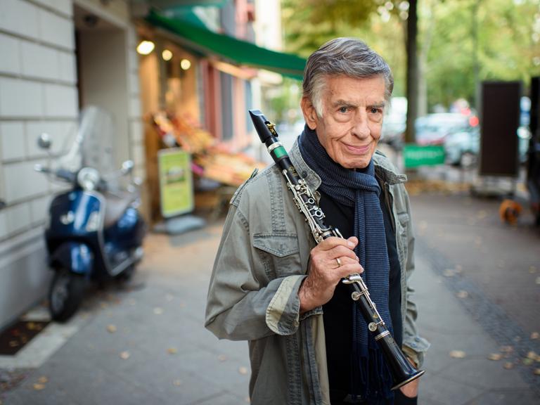 Rolf Kühn, Jazzmusiker und Komponist, steht im Berliner Bezirk Charlottenburg auf einem Gehweg und hält seine Klarinette in der Hand. Er hat graue, zur Seite gekämmte Haare und trägt einen blauen Schal um den Hals.