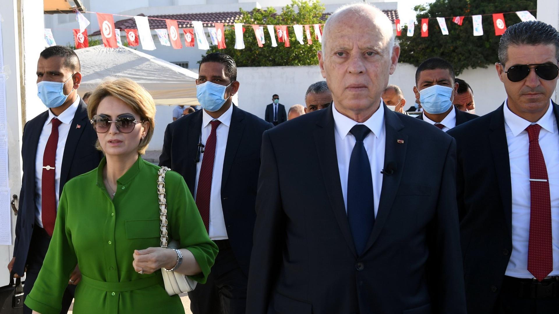 Tunesiens Präsident Kais Saied, begleitet von seiner Ehefrau und mehreren Leibwächtern, geht durch eine mit Wimpeln geschmückte Straße auf die Kamera zu. Tunesiens Präsident Kais Saied, begleitet von seiner Ehefrau und mehreren Leibwächtern, geht durch eine mit Wimpeln geschmückte Straße auf die Kamera zu.