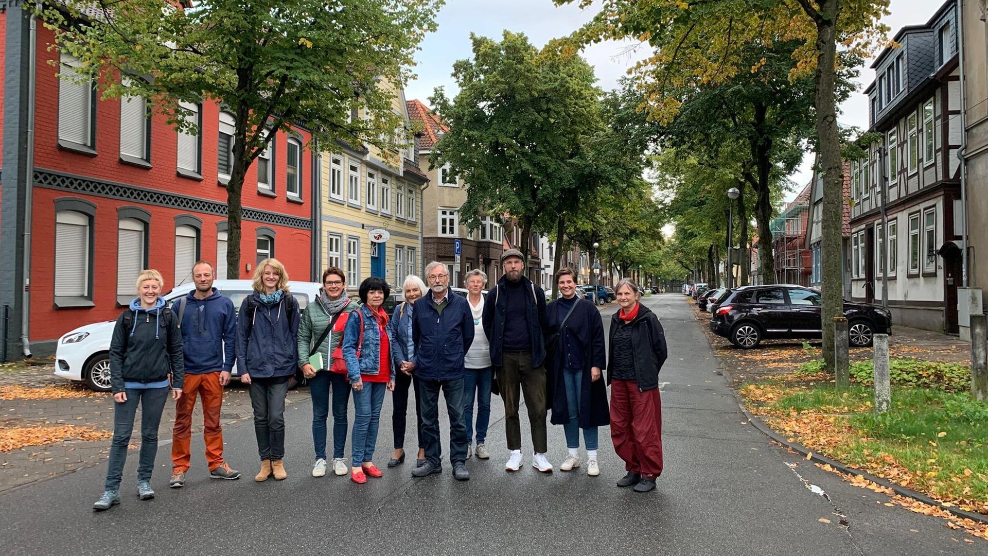 Zehn Menschen stehen nebeneinander mitten auf der Straße und schauen in die Kamera. Im Hintergrund ist eine Straße zu sehen, an deren Rändern Autos geparkt sind und Linden stehen. Zehn Menschen stehen nebeneinander mitten auf der Straße und schauen in die Kamera. Im Hintergrund ist eine Straße zu sehen, an deren Rändern Autos geparkt sind und Linden stehen.
