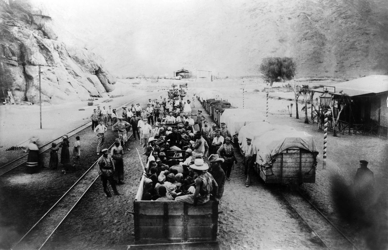 Historisches Schwarzweißfoto eines Bahnhofs, an dem Gefangene von Soldaten bewacht in offenen Eisenbahnwaggons sitzen. Historisches Schwarzweißfoto eines Bahnhofs, an dem Gefangene von Soldaten bewacht in offenen Eisenbahnwaggons sitzen.