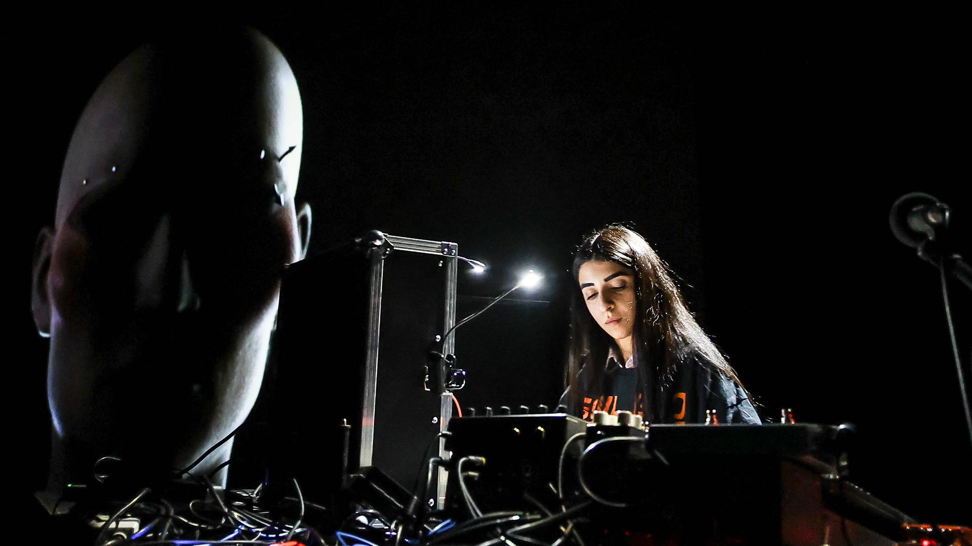 Die iranisch-deutsche Künstlerin Nazanin Noori steht bei einem Konzert beim CTM-Festival 2023 an technischen Geräten. Daneben ist ein projiziertes Gesicht zu sehen, ein Werk der Künstlerin Diana Azzuz, die einen ukrainisch-syrischen Hintergrund hat. Die iranisch-deutsche Künstlerin Nazanin Noori steht bei einem Konzert beim CTM-Festival 2023 an technischen Geräten. Daneben ist ein projiziertes Gesicht zu sehen, ein Werk der Künstlerin Diana Azzuz, die einen ukrainisch-syrischen Hintergrund hat.
