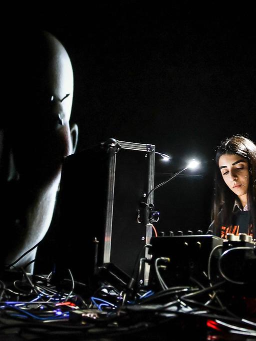 Die iranisch-deutsche Künstlerin Nazanin Noori steht bei einem Konzert beim CTM-Festival 2023 an technischen Geräten. Daneben ist ein projiziertes Gesicht zu sehen, ein Werk der Künstlerin Diana Azzuz, die einen ukrainisch-syrischen Hintergrund hat. 