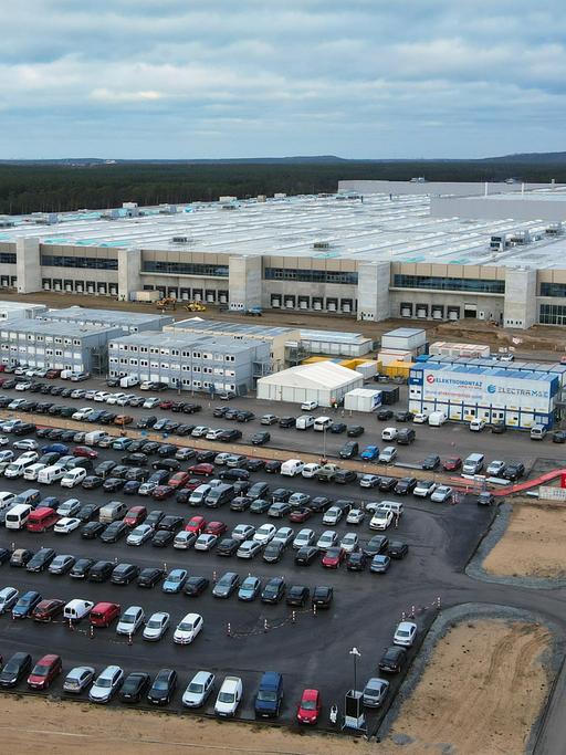 Die Lauftaufnahme mit einer Drohne zeigt eine große Baustelle. Im Vordergrund sind über hundert Autos geparkt, dann kommen Container, dahinter erhebt sich das Fabrikgebäude.