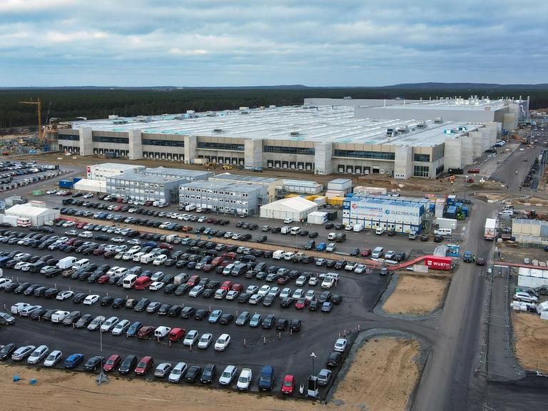 Die Lauftaufnahme mit einer Drohne zeigt eine große Baustelle. Im Vordergrund sind über hundert Autos geparkt, dann kommen Container, dahinter erhebt sich das Fabrikgebäude.