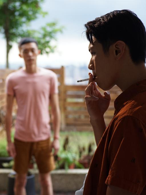 Ein Mann lehnt an einem Türrahmen und zieht cool an einer Zigarette. Im Garten dahinter steht ein weiterer Mann im Unschärfebereich des Bildes und schaut den rauchenden Mann an.