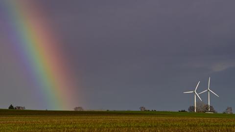 Zwei Windenergieanlagen stehen auf einem Feld, während sich daneben ein Regenbogen vor dunklen Himmel abzeichnet. 