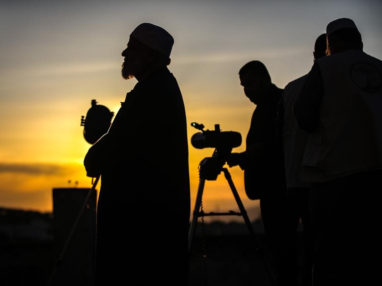 Die Silhouetten syrischer Gelehrter, die mit einem Teleskop den Mond beobachten, zeichnen sich gegen den rötlich schimmernden Himmel ab.