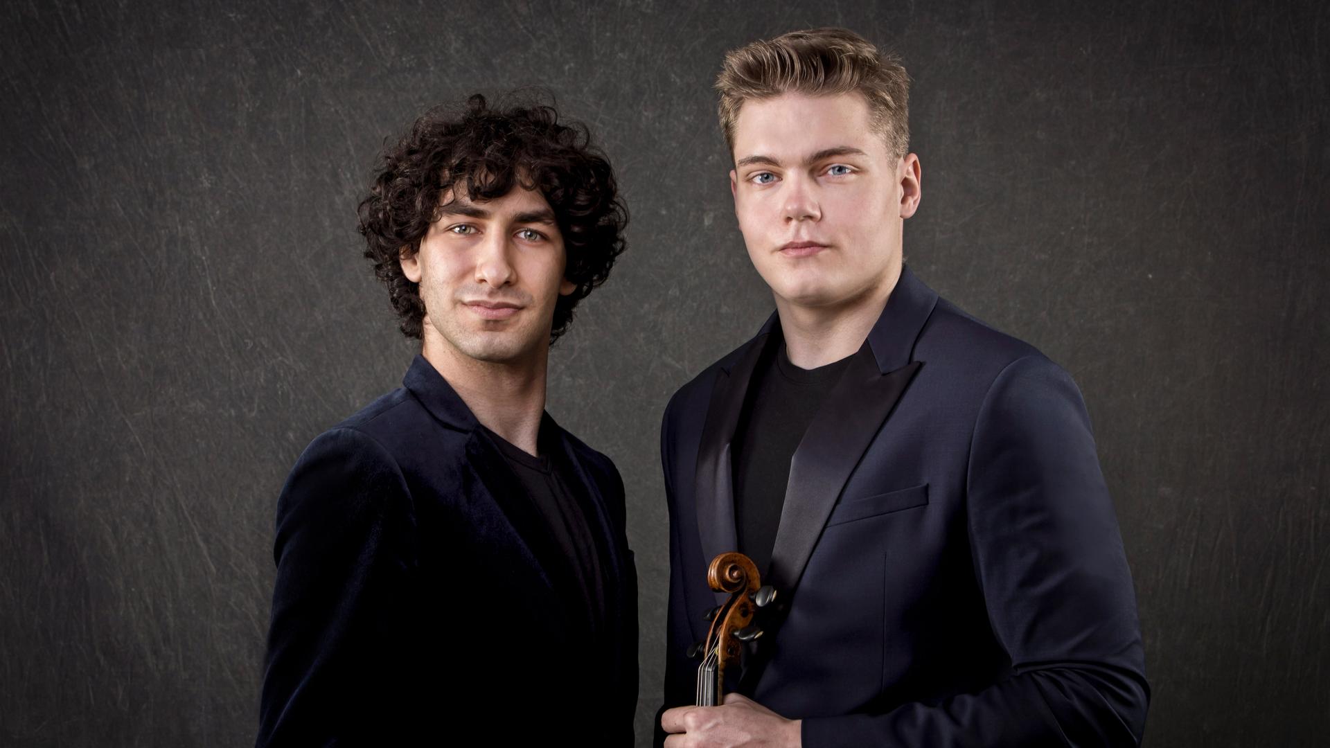 Zwei junge Männer in Konzertkleidung blicken in die Kamera. Der Mann auf der rechten Seite hält seine Violine vor dem Körper.