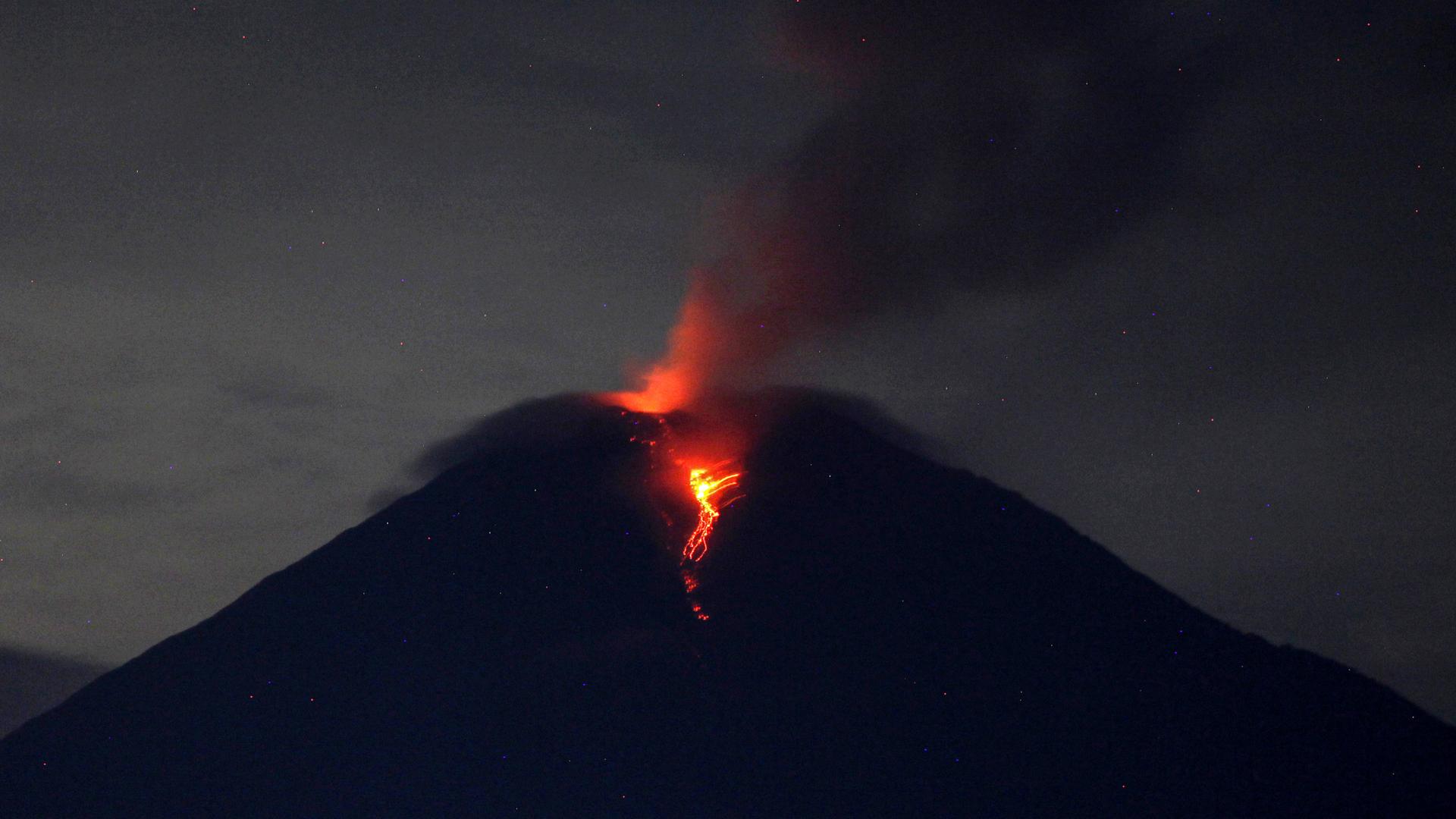 Blick aus der Ferne auf die Spitze des Vulkans im Halbdunkel, aus dem rote Lava strömt.