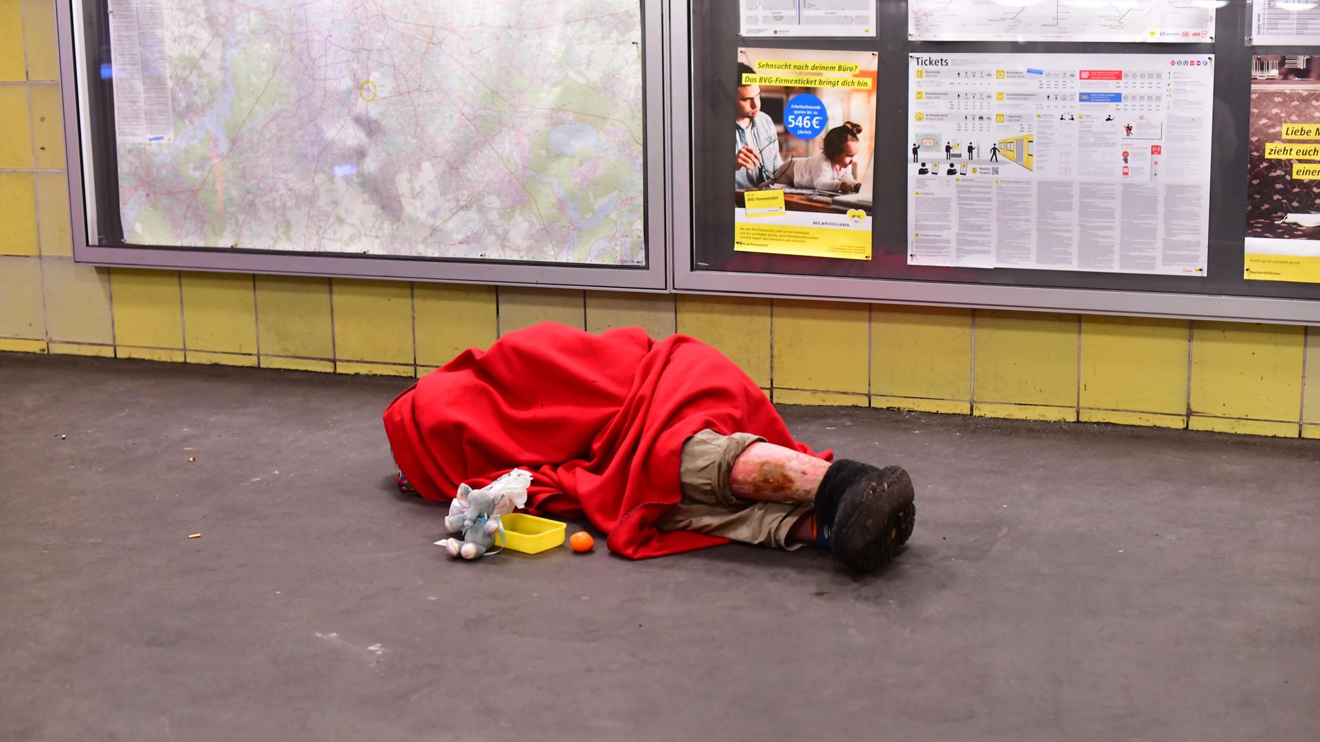 Ein Obdachloser liegt in einen Schlafsack gehüllt morgens im Bahnhof Tempelhof mit einem Becher für Spenden vor sich und schläft auf dem Boden.