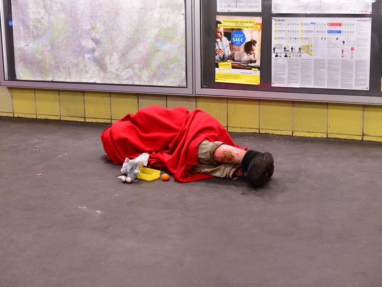 Ein Obdachloser liegt in einen Schlafsack gehüllt morgens im Bahnhof Tempelhof mit einem Becher für Spenden vor sich und schläft auf dem Boden.