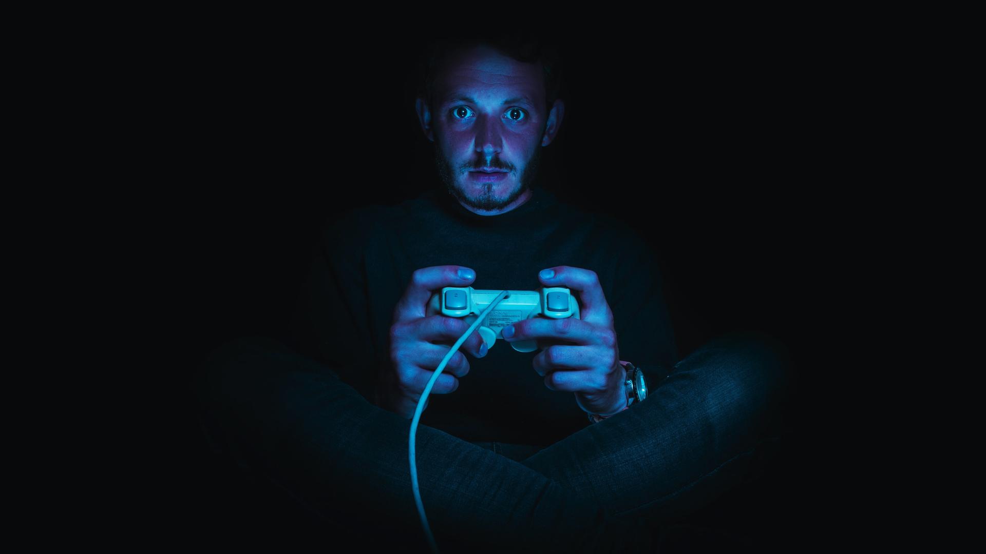 Ein Mann sitzt in einer dunklen Umgebung und hält einen Gaming-Controller. Seine Gesichtshaut ist rötlich, seine Augen sind weit aufgerissen.
