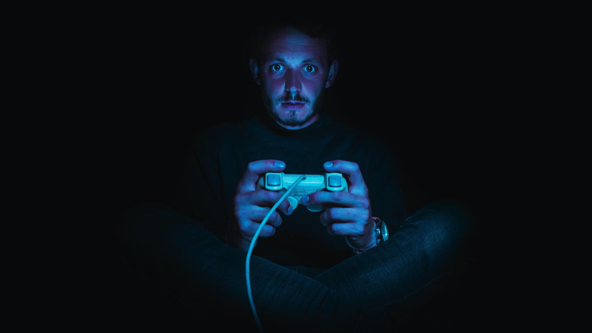 Ein Mann sitzt in einer dunklen Umgebung und hält einen Gaming-Controller. Seine Gesichtshaut ist rötlich, seine Augen sind weit aufgerissen. Ein Mann sitzt in einer dunklen Umgebung und hält einen Gaming-Controller. Seine Gesichtshaut ist rötlich, seine Augen sind weit aufgerissen.