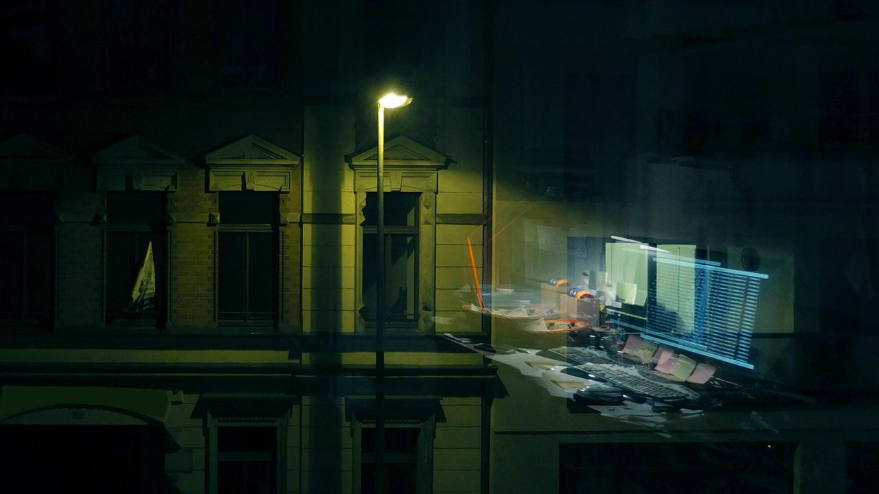 Computermonitore, die sich in einer Fensterscheibe spiegeln. Draußen leuchtet eine Straßenlaterne.
