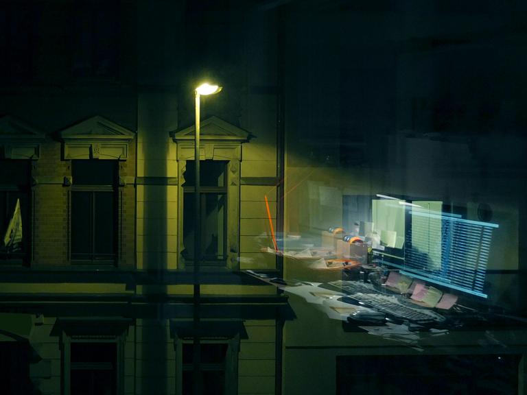 Computermonitore, die sich in einer Fensterscheibe spiegeln. Draußen leuchtet eine Straßenlaterne.