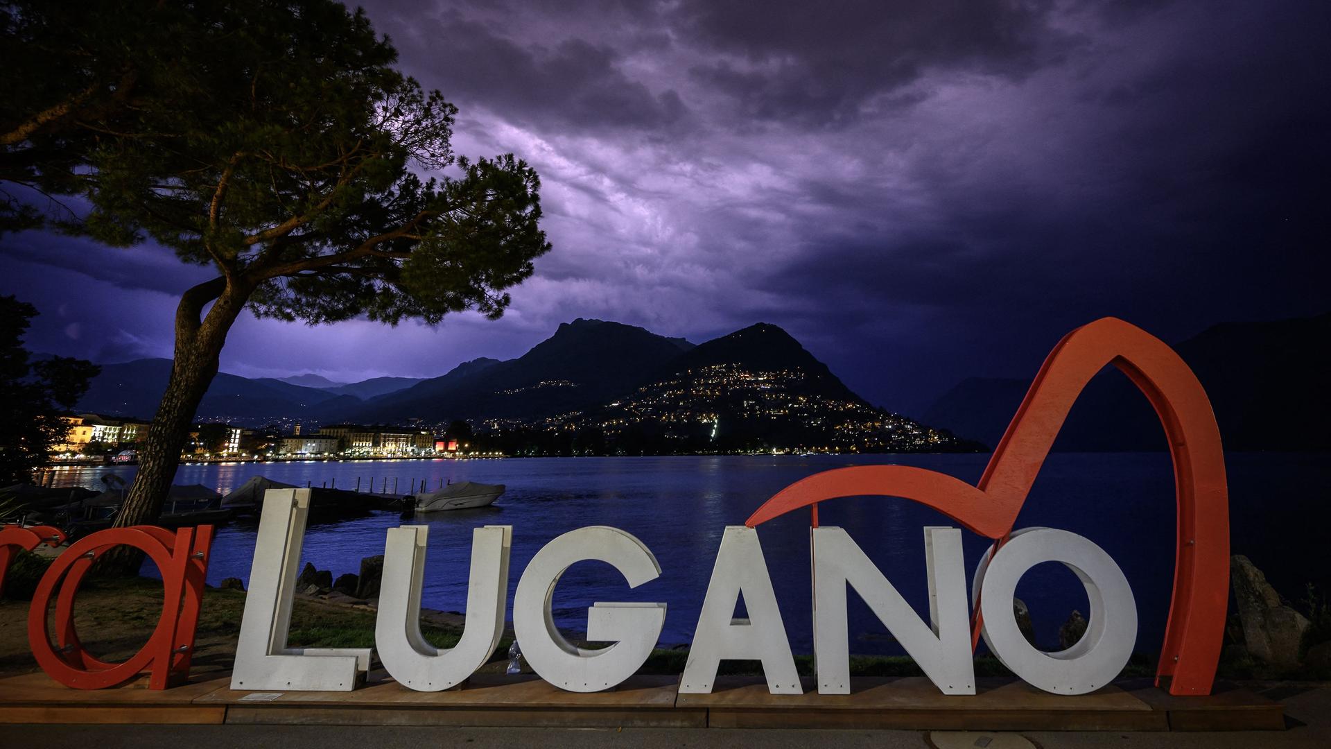 Das Logo der Konferenz in Lugano zum Wiederaufbau der kriegszerstörten Gebiete in der Ukraine vor dunklem Himmel und einem See.