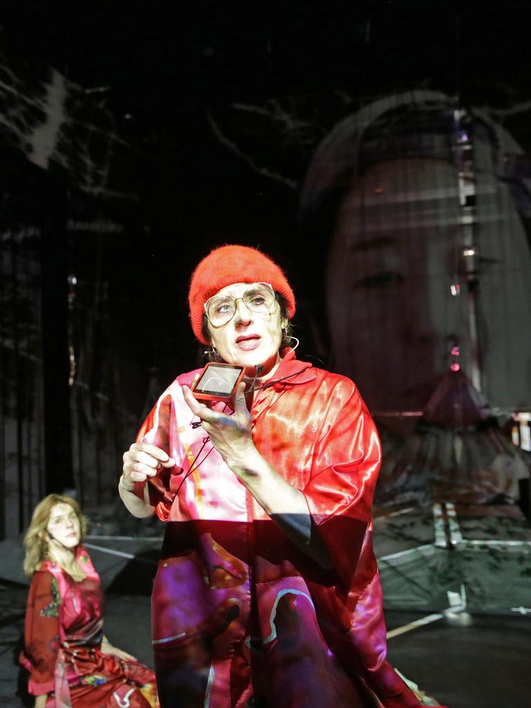 Szenenbild aus der Aufführung von "Mauern" des Künstler*innenkollektivs "She She Pop". Zu sehen sind verschiedene Überblendungen. Im Vordergrund eine Person mit roter Mütze, die einen Spiegel hält. Links davon, knieend, eine Frau im roten Kleid. Rechts im Hintergrund ein eingeblendetes Gesicht. 
