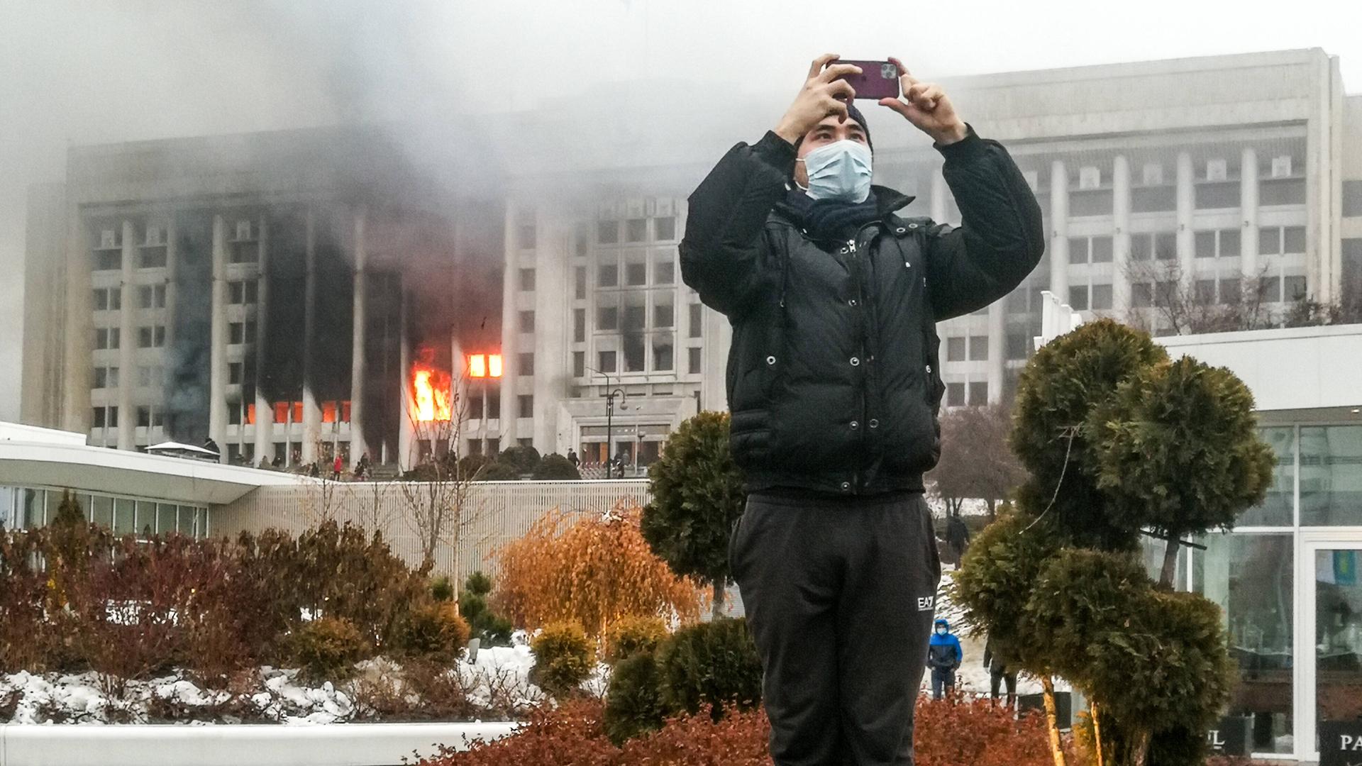 Ein junger Mann macht in der kasachischen Stadt Almaty mit seinem Smartphone ein Foto, im Hintergrund brennt das Amtsgebäude des Bürgermeisters Ein junger Mann macht in der kasachischen Stadt Almaty mit seinem Smartphone ein Foto, im Hintergrund brennt das Amtsgebäude des Bürgermeisters
