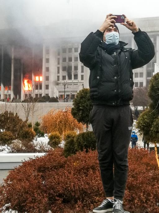 Ein junger Mann macht in der kasachischen Stadt Almaty mit seinem Smartphone ein Foto, im Hintergrund brennt das Amtsgebäude des Bürgermeisters