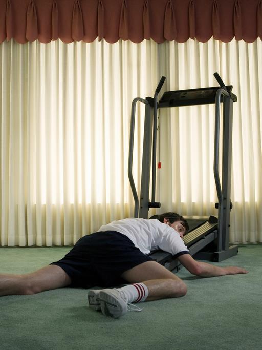 Ein junger Mann hängt erschöpft am Boden vor einem Fitness Laufband.