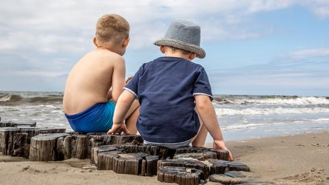 Das Foto zeigt zwei kleine Kinder in Rückenansicht:. Sie  sitzen auf Holzpfählen am Strand. Ein Kind trägt einen Sonnenhut. Strand und Meer sind leer.